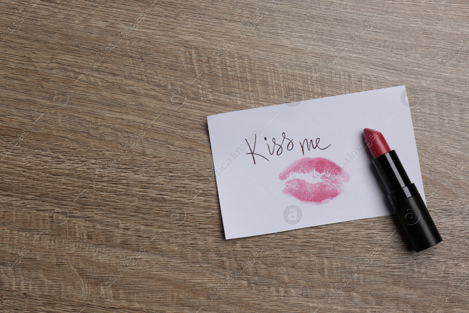 Photo of Paper card with lip print, phrase Kiss Me and lipstick on wooden table, top view. Space for text