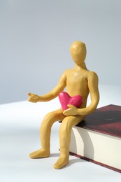 Photo of Yellow plasticine human figure with pink heart sitting on book against white background