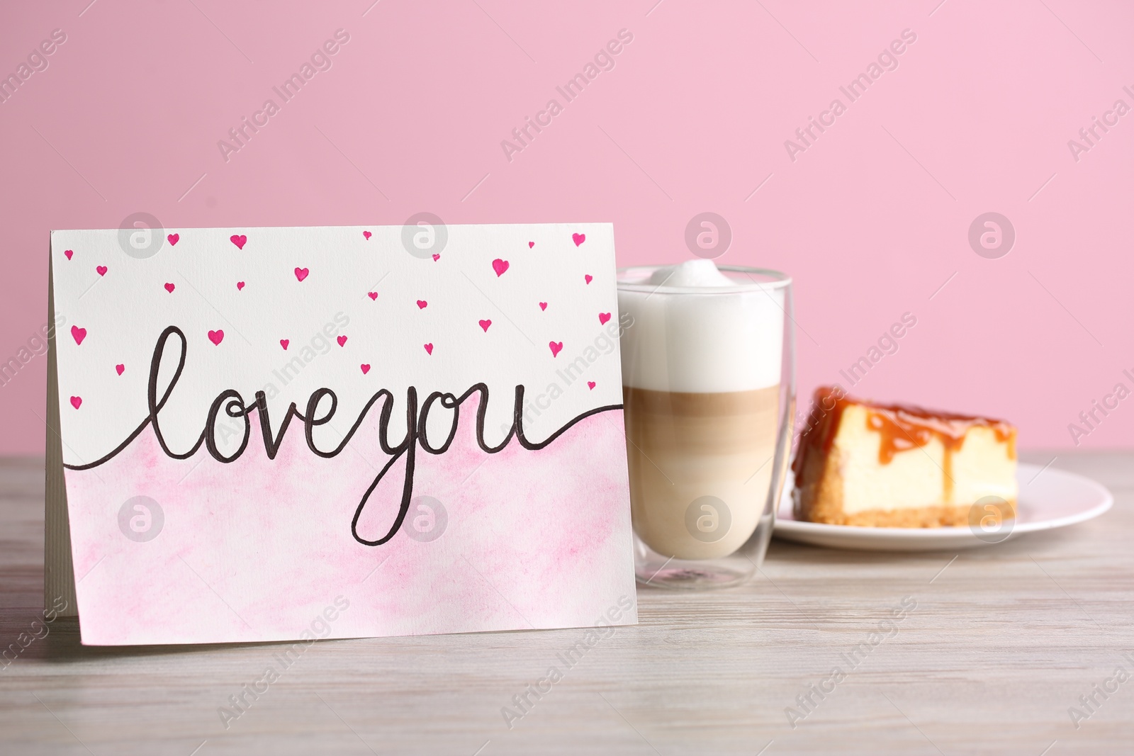 Photo of Card with phrase Love You, latte macchiato and cake on white wooden table against pink wall