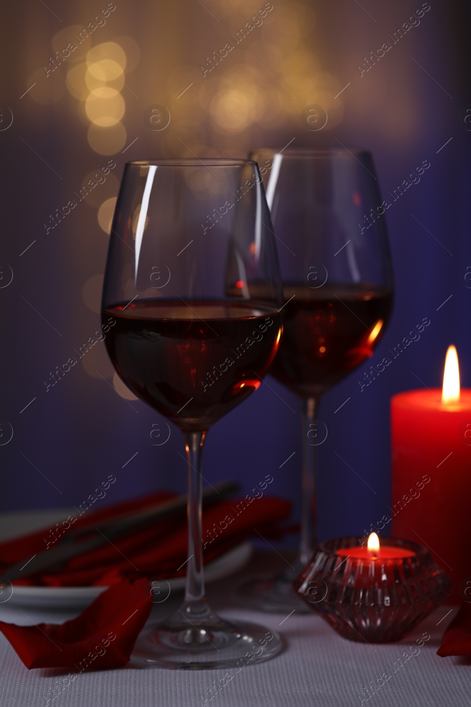 Photo of Romantic table setting with glasses of red wine and burning candles on grey table against blurred lights