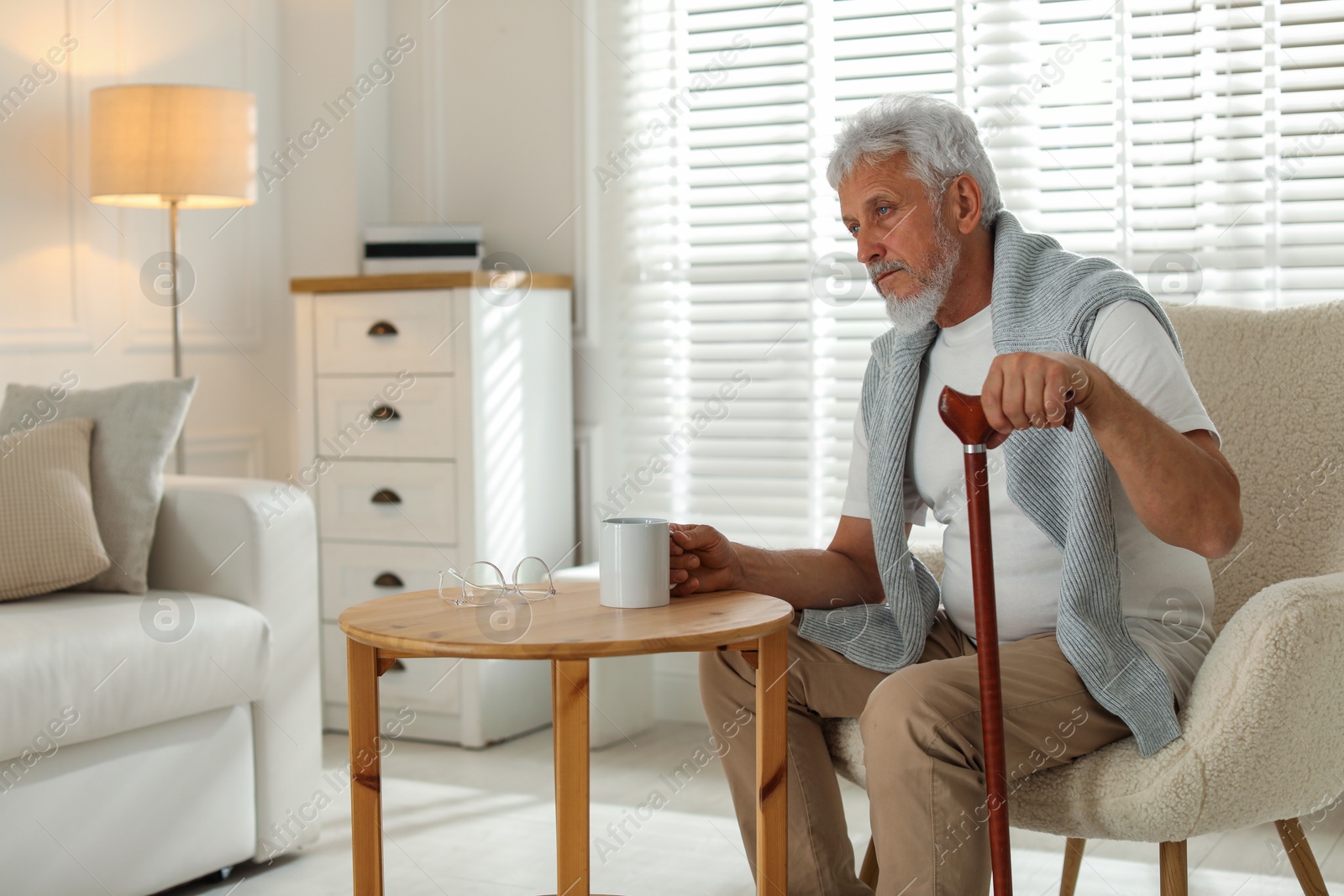 Photo of Lonely senior man with walking cane and cup of drink sitting at table indoors