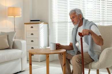 Photo of Lonely senior man with walking cane and cup of drink sitting at table indoors