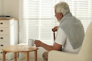 Photo of Lonely senior man with walking cane and cup of drink sitting on sofa at home