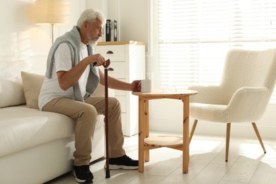 Photo of Lonely senior man with walking cane and cup of drink sitting on sofa at home