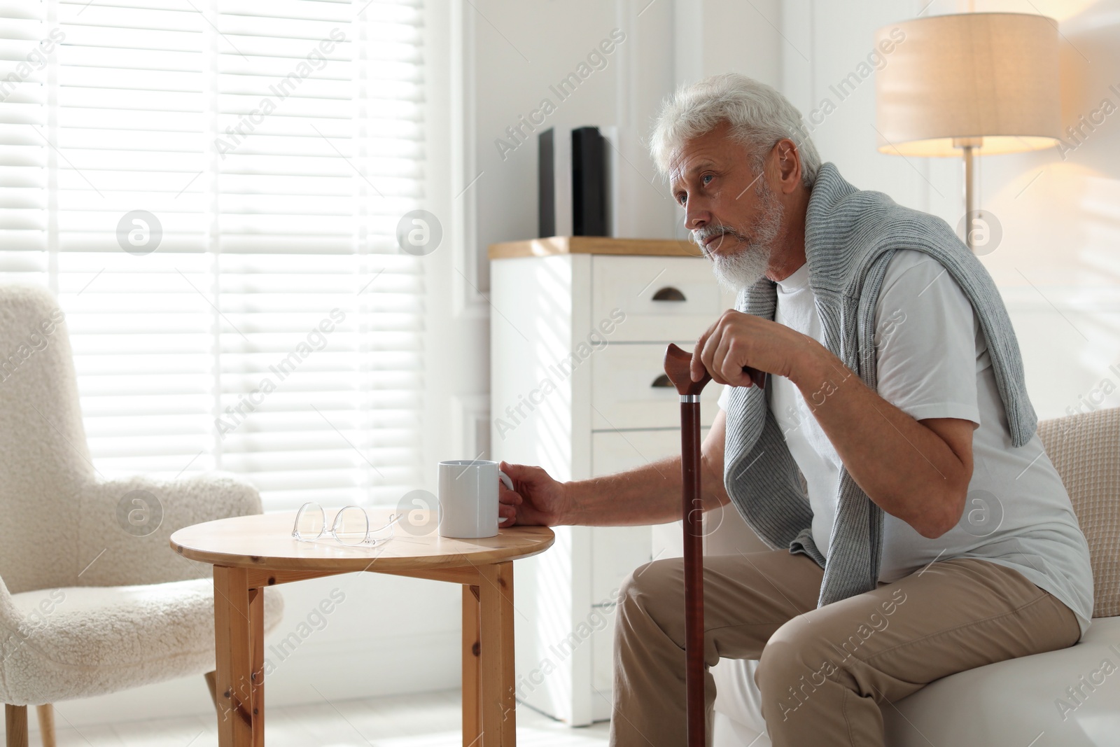 Photo of Lonely senior man with walking cane and cup of drink sitting on sofa at home