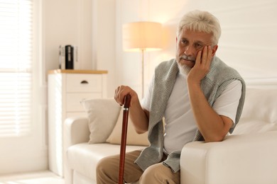 Lonely senior man with walking cane sitting on sofa at home. Space for text