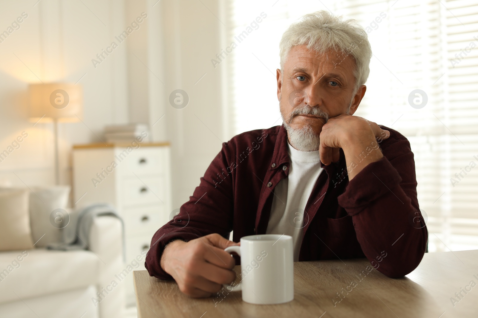 Photo of Lonely senior man with cup of drink sitting at wooden table in room