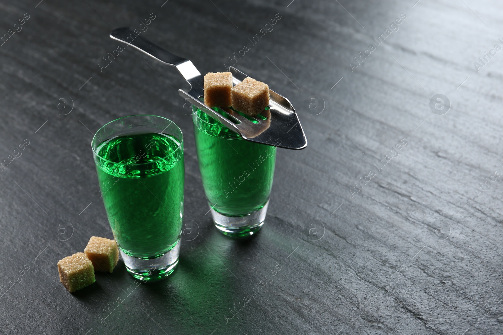 Photo of Absinthe in shot glasses, brown sugar and slotted spoon on black table, closeup. Space for text