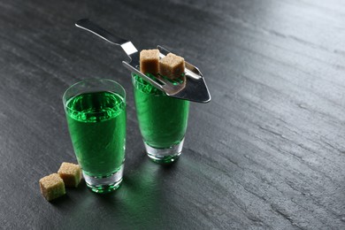 Photo of Absinthe in shot glasses, brown sugar and slotted spoon on black table, closeup. Space for text