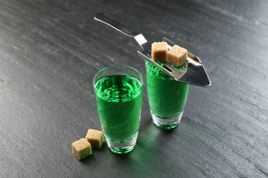 Photo of Absinthe in shot glasses, brown sugar and slotted spoon on black table, closeup