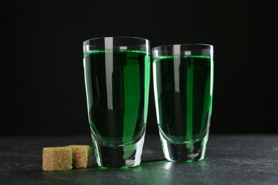 Photo of Absinthe in shot glasses and brown sugar on table against black background, closeup