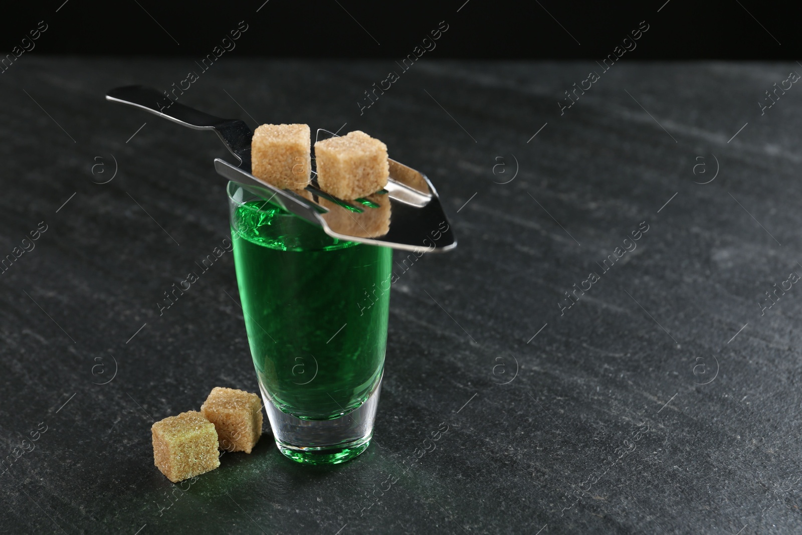 Photo of Absinthe in shot glass, brown sugar and slotted spoon on black table, closeup. Space for text