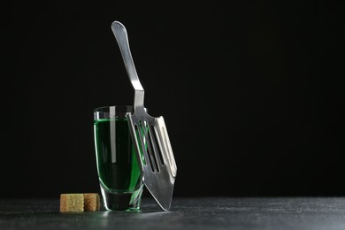 Photo of Absinthe in shot glass, brown sugar and slotted spoon on table against black background, space for text