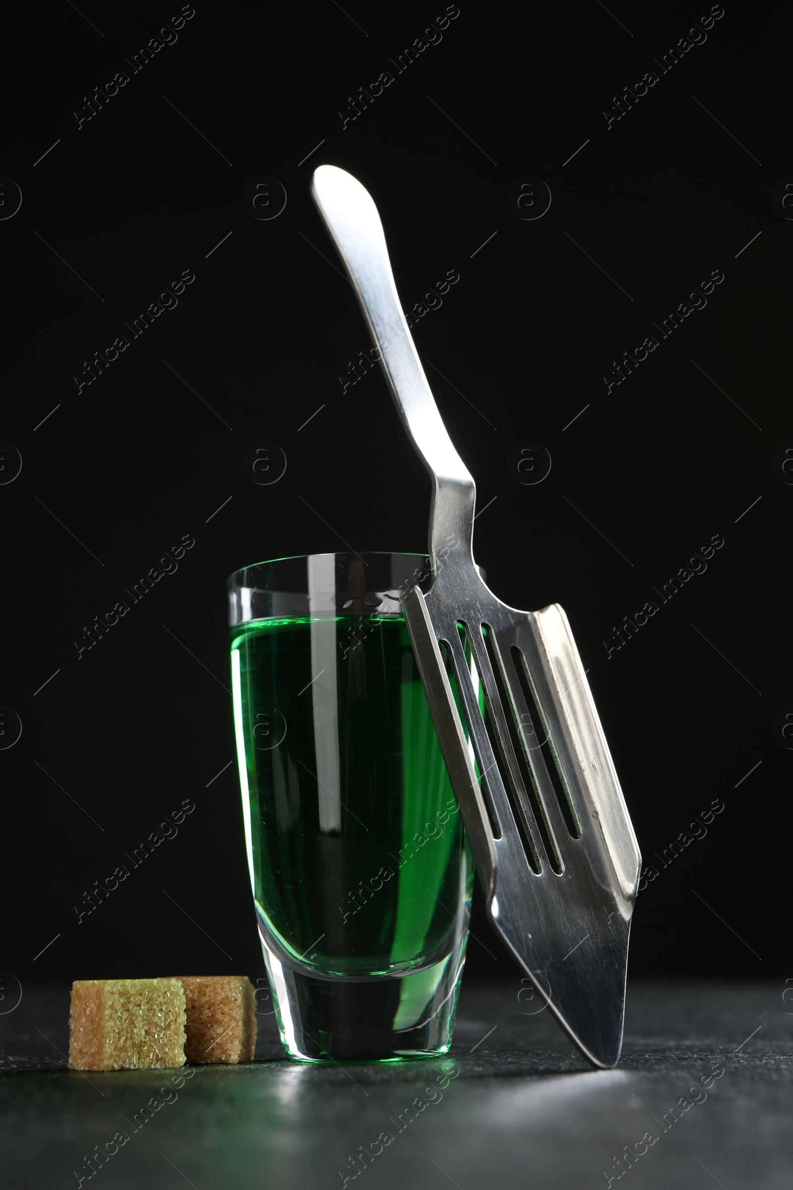 Photo of Absinthe in shot glass, brown sugar and slotted spoon on table against black background