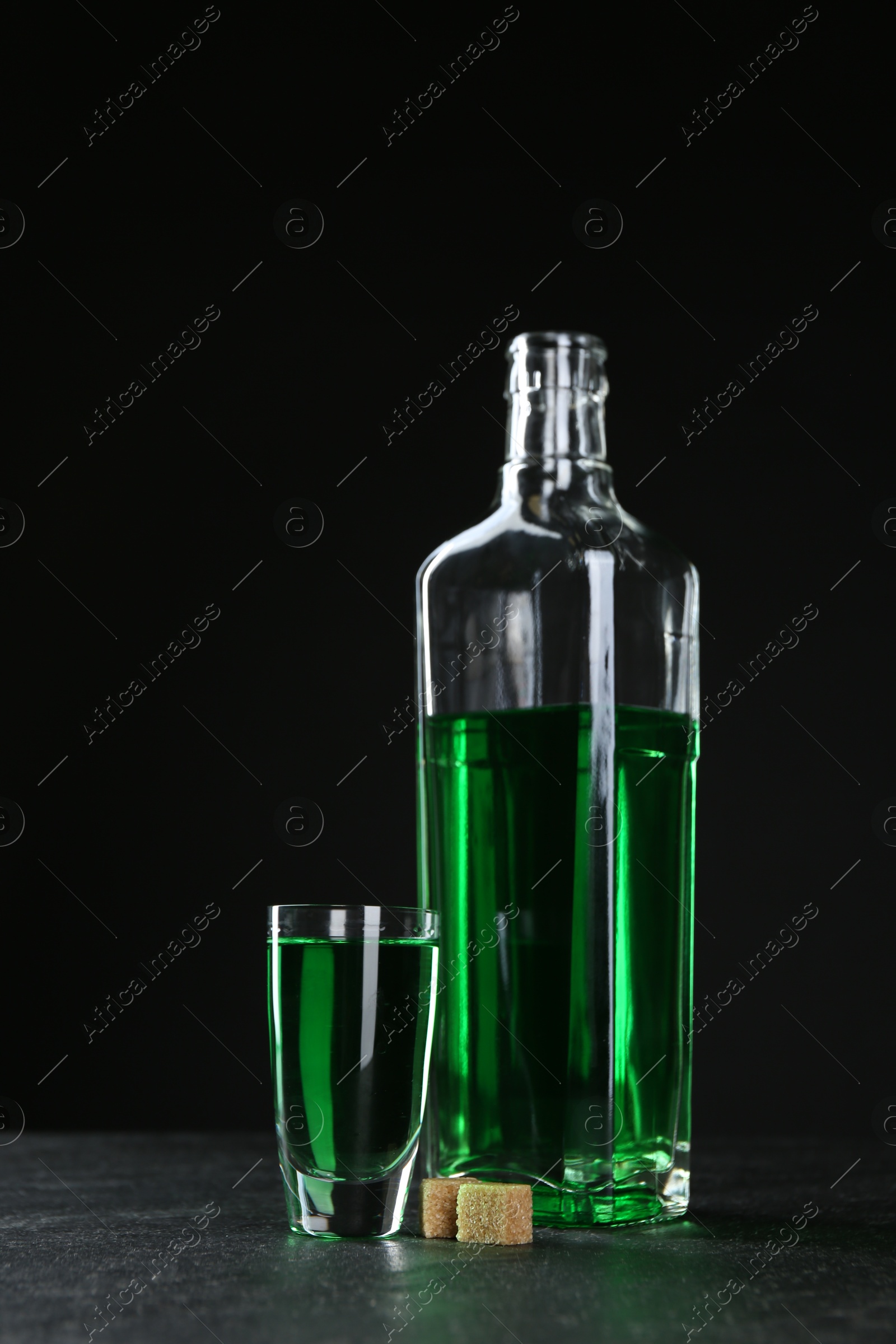 Photo of Absinthe in shot glass, bottle and brown sugar on table against black background