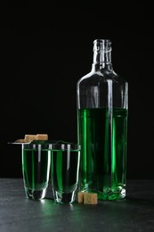 Photo of Absinthe in shot glasses, bottle, brown sugar and slotted spoon on table against black background