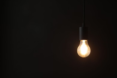 Photo of Glowing light bulb hanging on dark background. Space for text