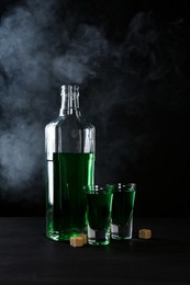 Photo of Absinthe in shot glasses, bottle and brown sugar on table against black background with smoke