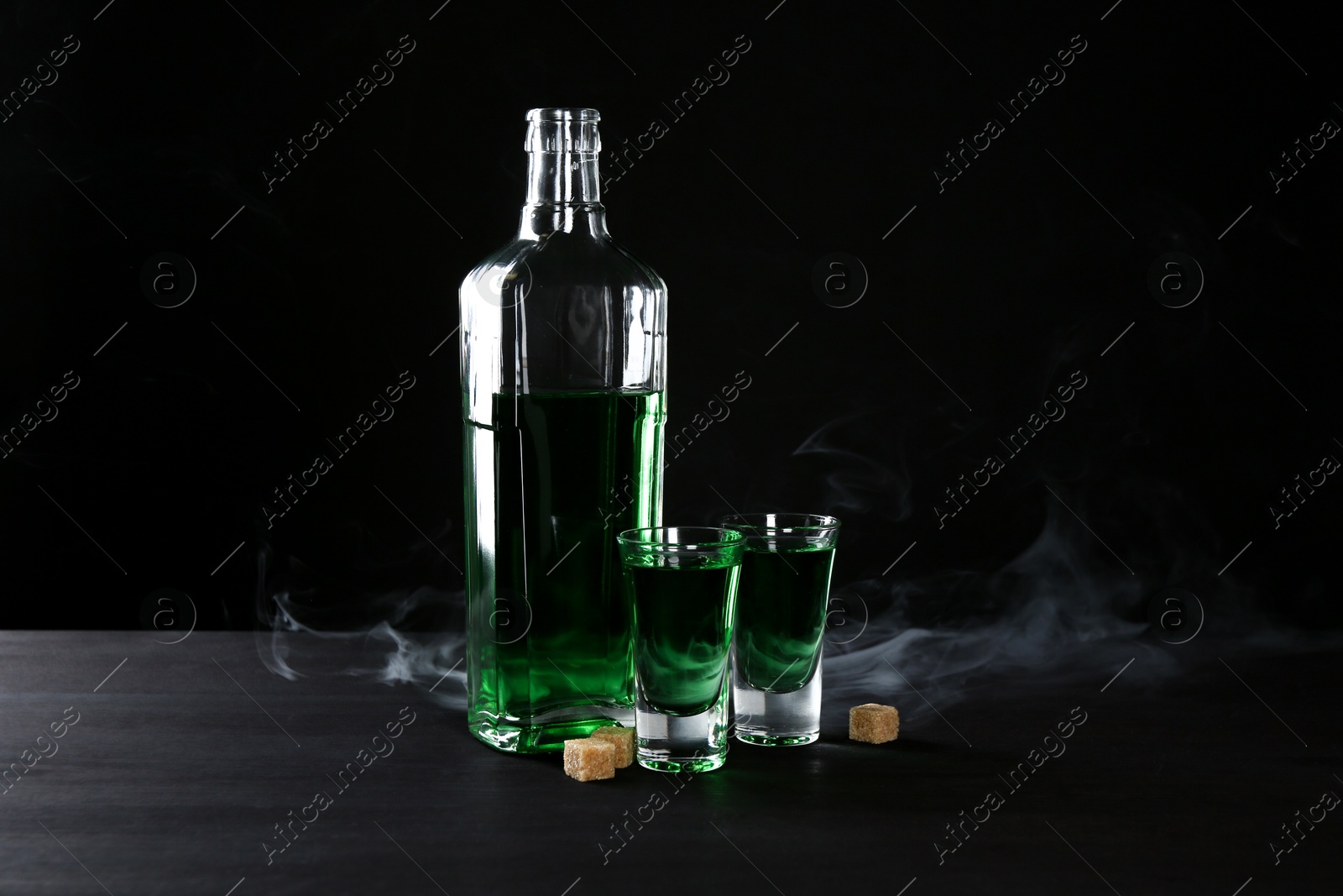 Photo of Absinthe in shot glasses, bottle and brown sugar on table against black background with smoke