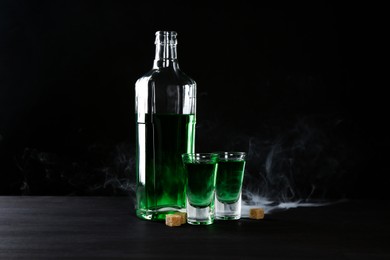 Photo of Absinthe in shot glasses, bottle and brown sugar on table against black background with smoke