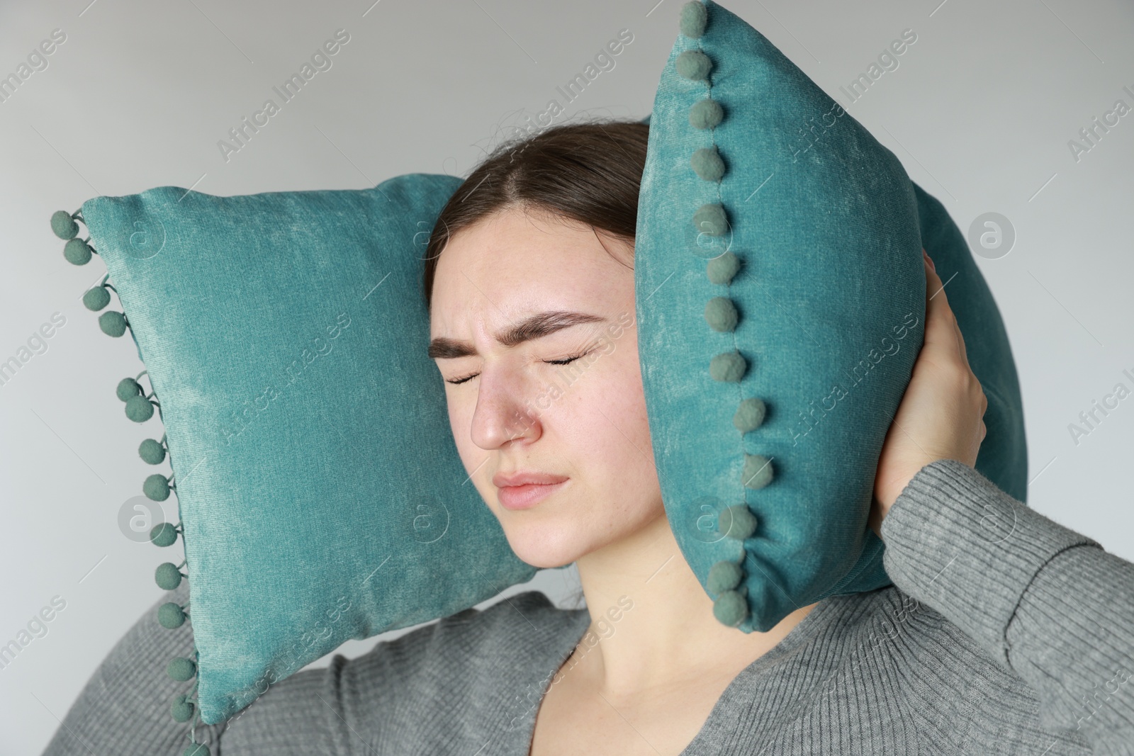 Photo of Distressed woman covering her ears with pillows from loud noise on light grey background