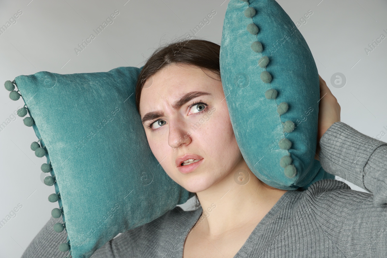 Photo of Distressed woman covering her ears with pillows from loud noise on light grey background