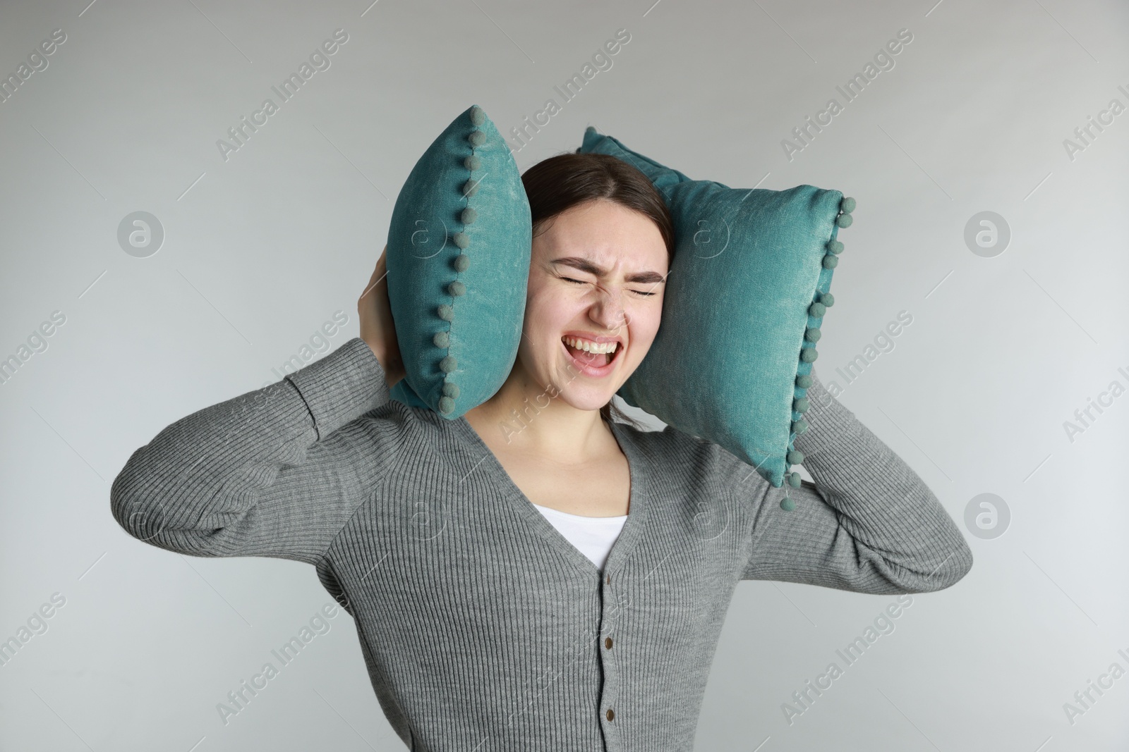 Photo of Distressed woman covering her ears with pillows from loud noise on light grey background