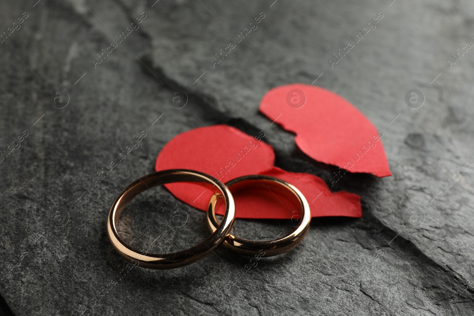 Photo of Halves of torn red paper heart and wedding rings on dark grey table. Broken heart