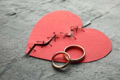 Photo of Broken heart. Torn red paper heart sewed with thread and wedding rings on grey table, closeup