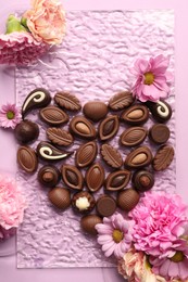 Heart made with delicious chocolate candies and beautiful flowers on pink background, flat lay