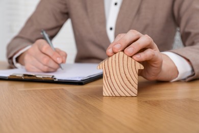 Photo of Property insurance. Real estate agent with house figure working at wooden table indoors, closeup