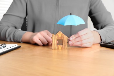 Photo of Property insurance. Real estate agent holding small umbrella above wooden house figure at table indoors, closeup