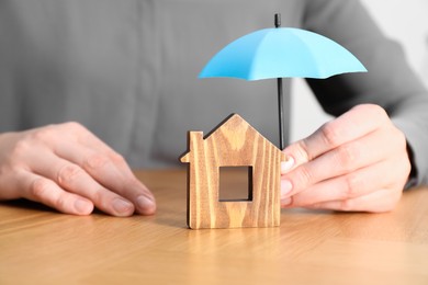 Photo of Property insurance. Real estate agent holding small umbrella above wooden house figure at table indoors, closeup