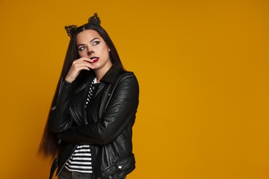 Photo of Woman with cat makeup and ears in leather jacket on orange background. Space for text