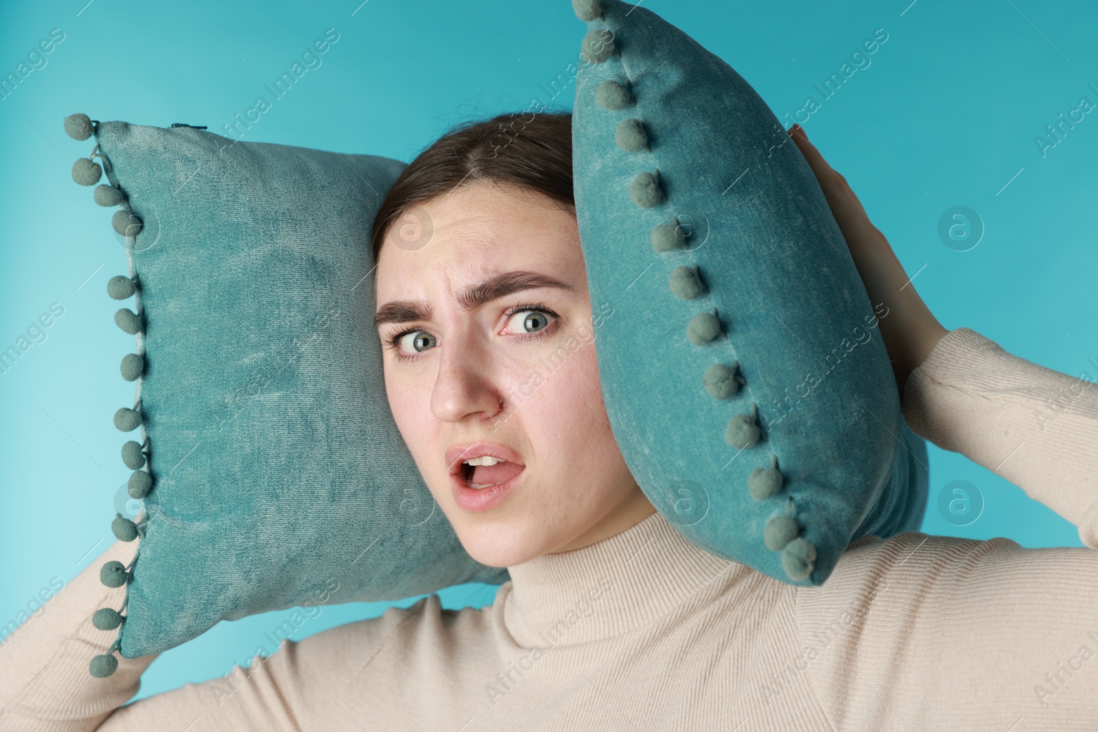 Photo of Distressed woman covering her ears with pillows from loud noise on blue background