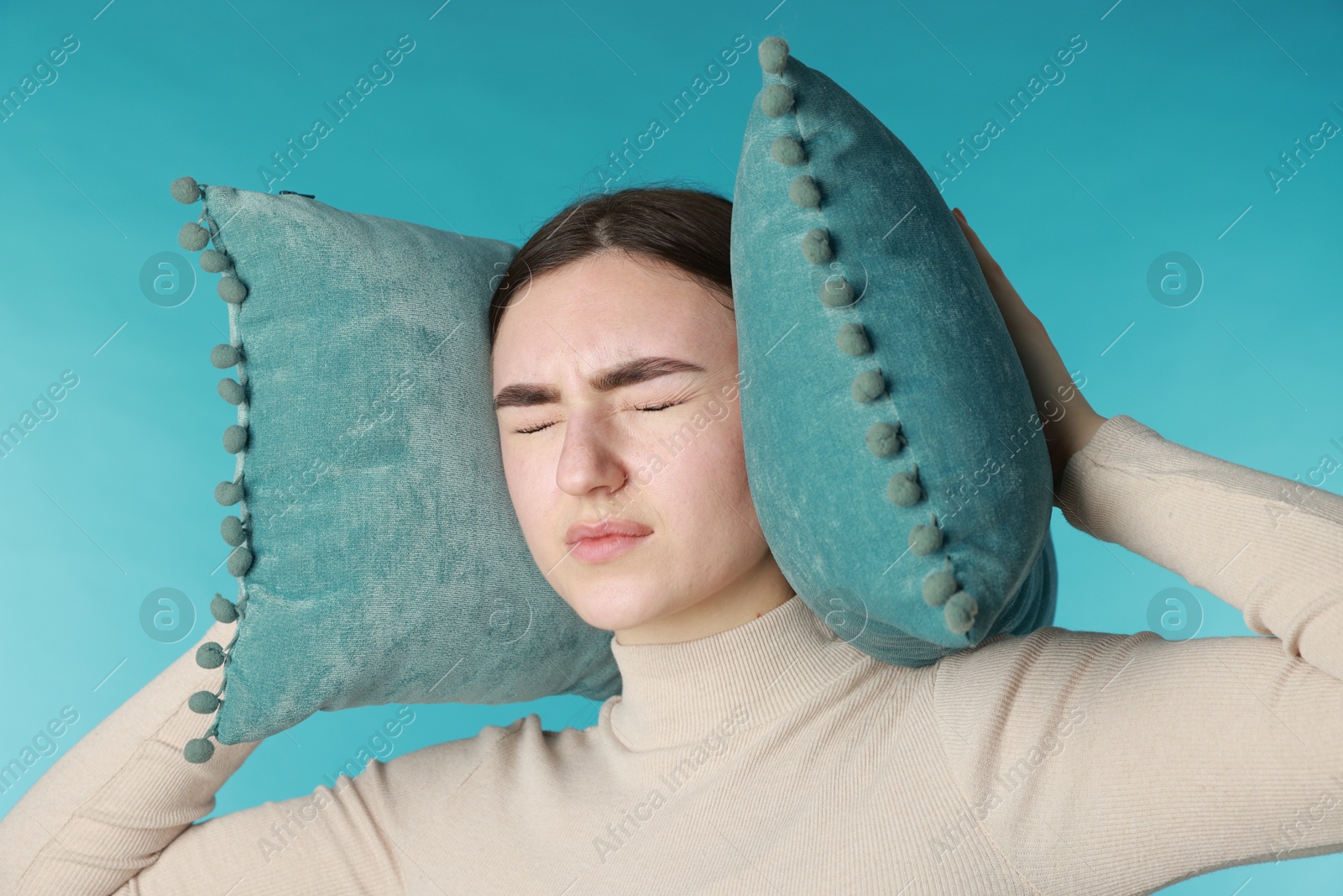 Photo of Distressed woman covering her ears with pillows from loud noise on blue background