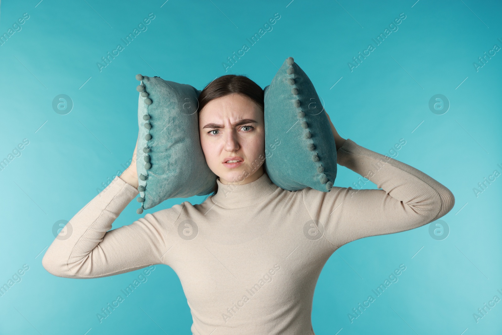 Photo of Frustrated woman covering her ears with pillows from loud noise on blue background