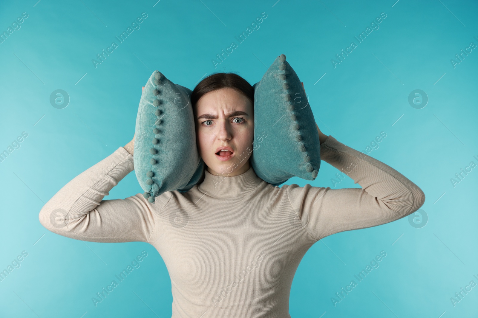 Photo of Frustrated woman covering her ears with pillows from loud noise on blue background