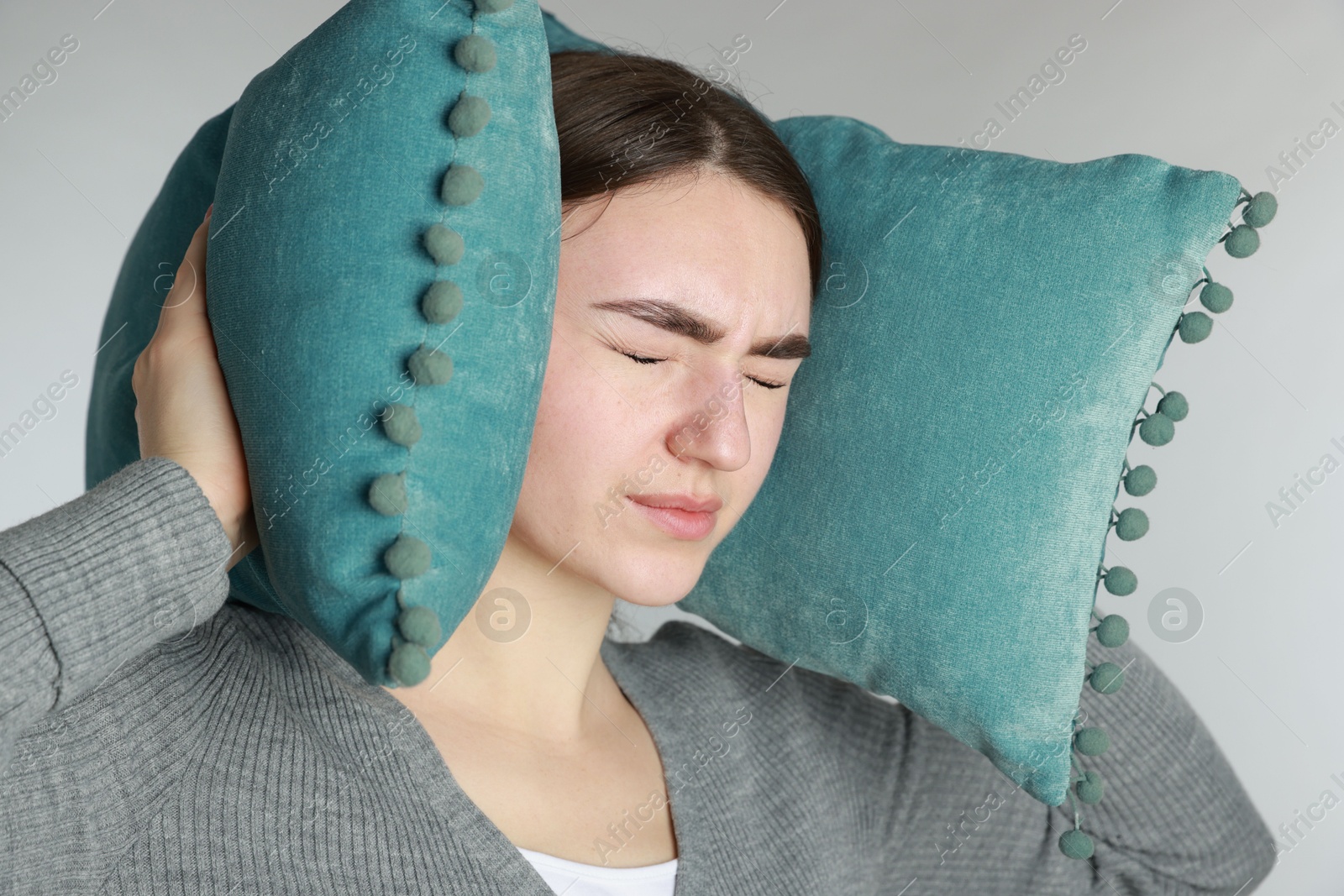 Photo of Distressed woman covering her ears with pillows from loud noise on light grey background