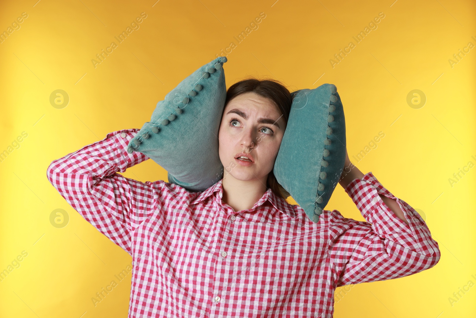 Photo of Distressed woman covering her ears with pillows from loud noise on yellow background