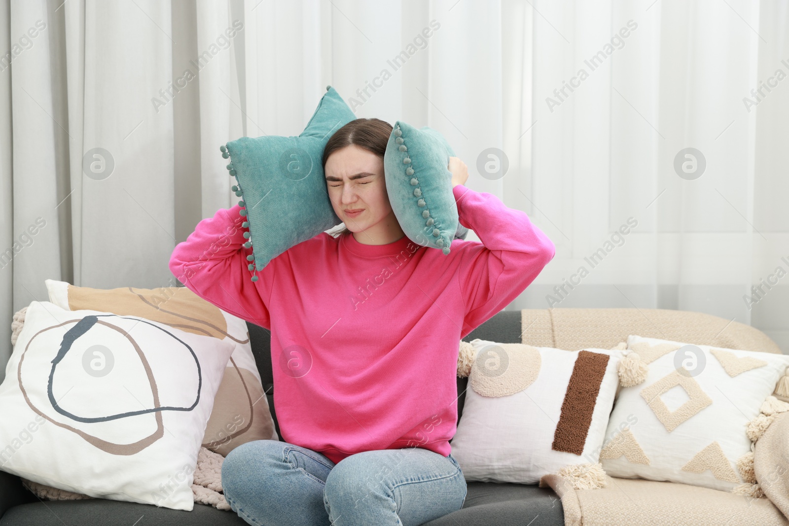 Photo of Annoyed woman covering her ears with pillows from loud noise at home