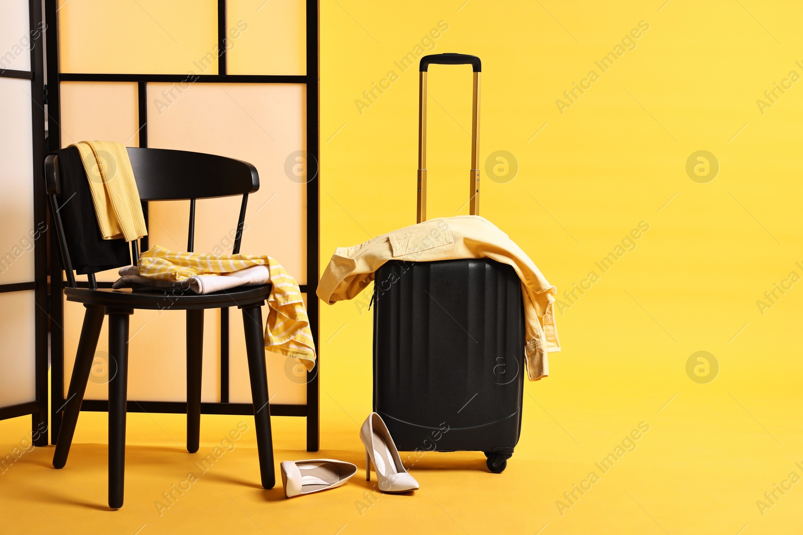 Suitcase with messy clothes and chair on yellow background Photo of Suitcase with messy clothes and chair on yellow background
