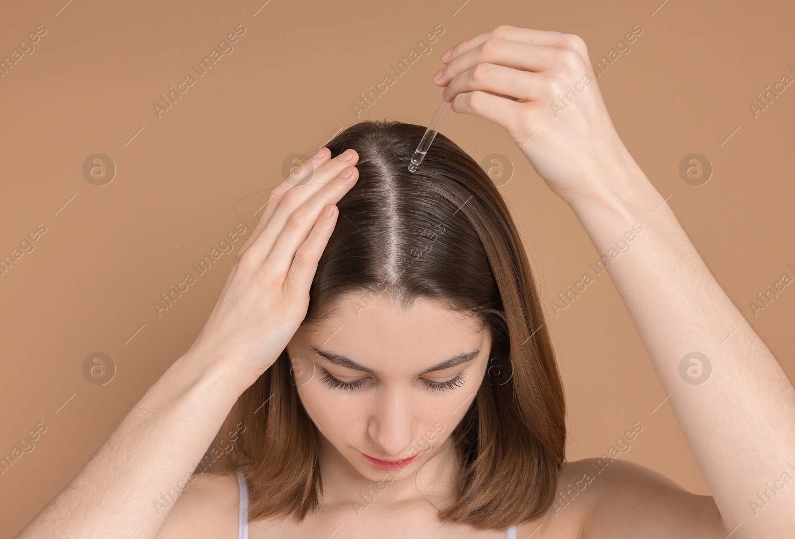 Photo of Hair loss problem. Girl applying serum onto hairline on dark beige background, closeup