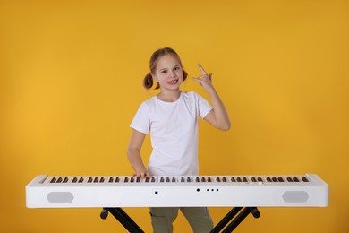 Photo of Cute girl playing synthesizer and showing rock gesture on orange background