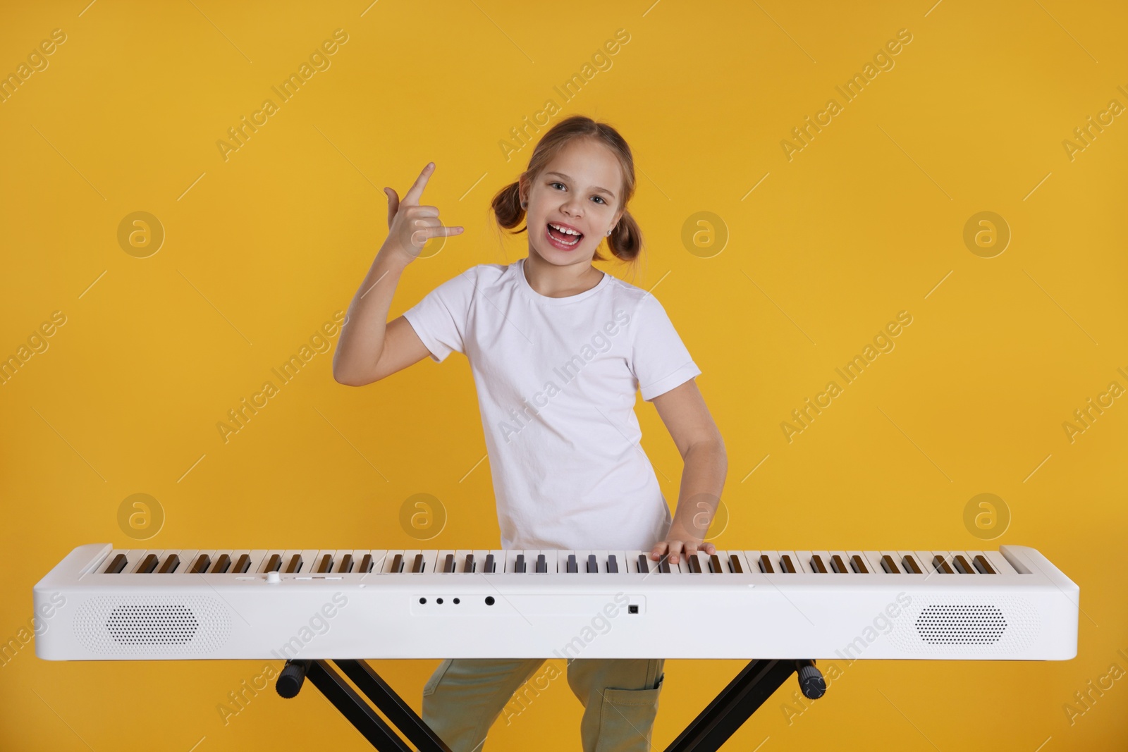 Photo of Cute girl playing synthesizer and showing rock gesture on orange background