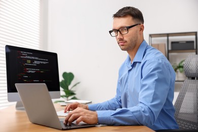 Photo of Programmer working on laptop and computer at wooden desk indoors