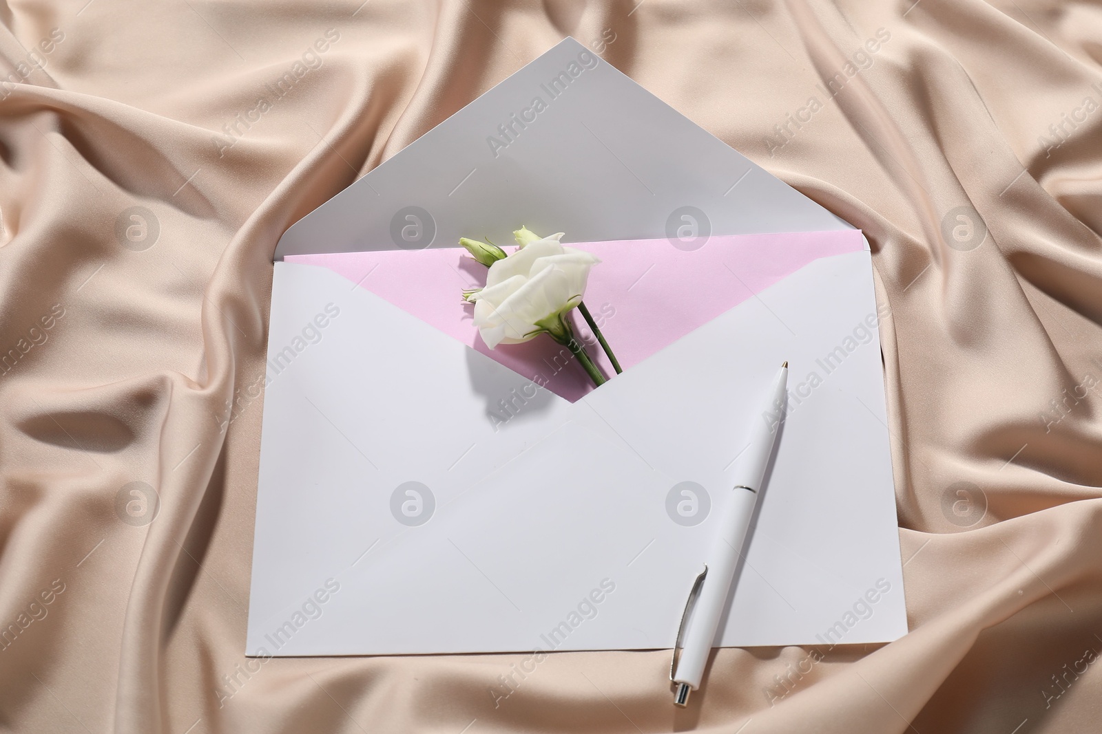 Photo of Paper envelope with letter, flower and pen on beige silk fabric, top view. Mockup for design