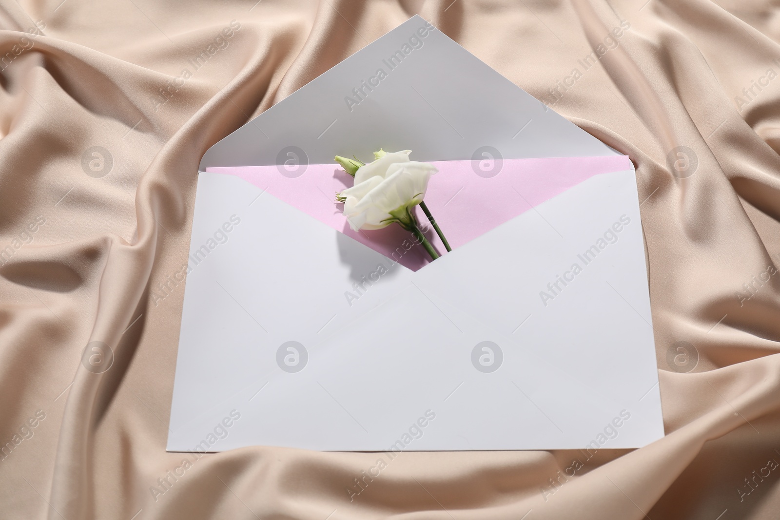 Photo of Paper envelope with letter and flower on beige silk fabric, above view. Mockup for design