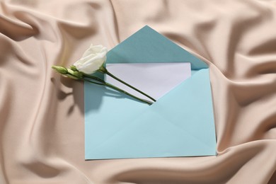 Photo of Paper envelope with letter and flowers on beige silk fabric, top view. Mockup for design