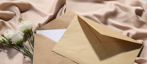 Photo of Kraft paper envelopes with letter and flowers on beige silk fabric, above view. Mockup for design